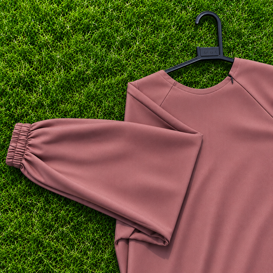 A dusty rose long-sleeve sweatshirt with an elasticized cuff, laid flat on a black hanger over green grass.