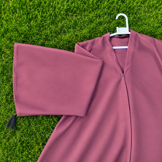 A dusty rose pink graduation gown laid flat on green grass, featuring a matching pleated sleeve with a black tassel and a white plastic hanger.