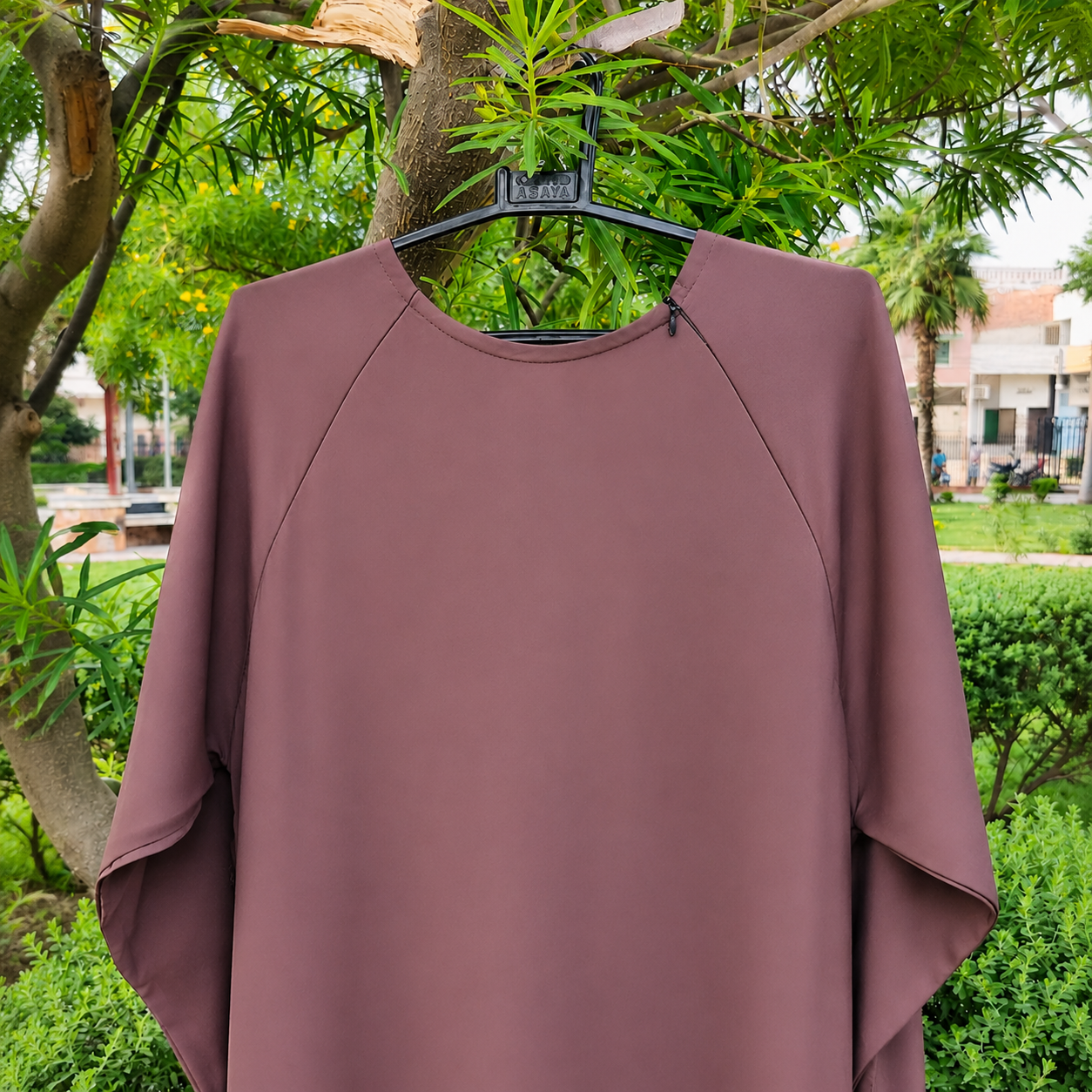 A loose-fitting, short-sleeve top in a muted mauve-brown color hanging on a black hanger outdoors against a background of green trees and shrubs.