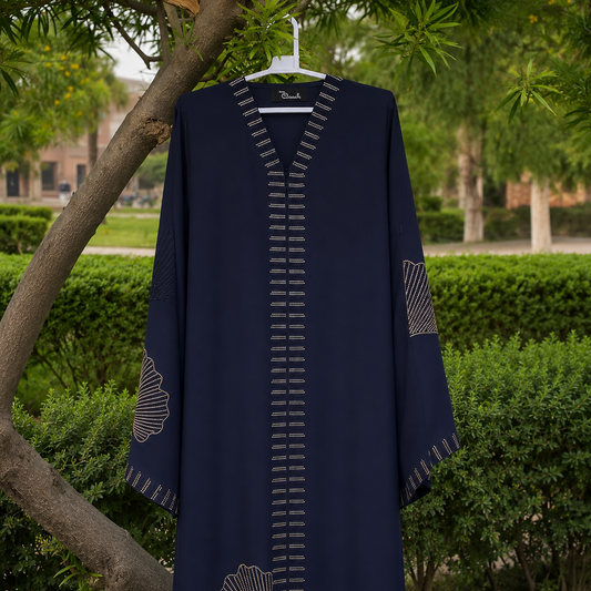 A navy blue abaya with geometric embroidered details and a V-neckline, hanging on a white hanger outdoors against a background of green hedges and trees.