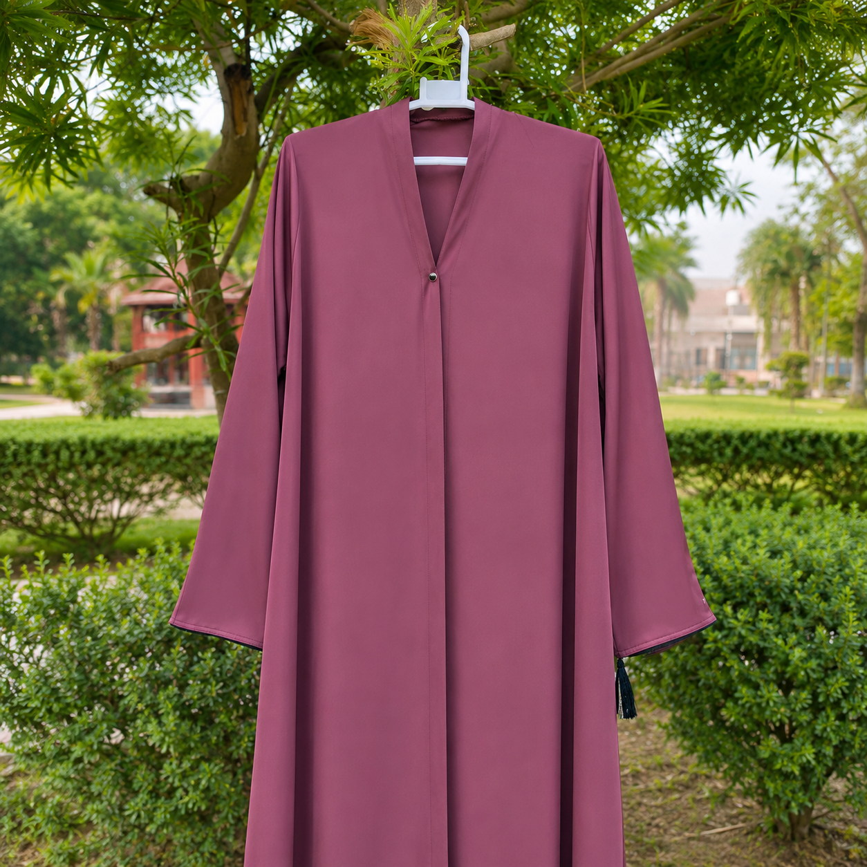 A long, flowing abaya dress in a dusty rose or mauve color, hanging on a white hanger outdoors against a background of green trees and bushes.