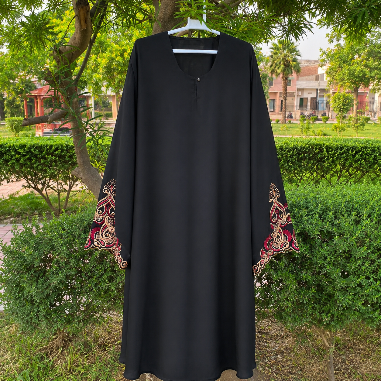 A black long-sleeve abaya dress with ornate red and gold embroidered patterns on the cuffs, hanging on a white hanger outdoors in front of green bushes and trees.