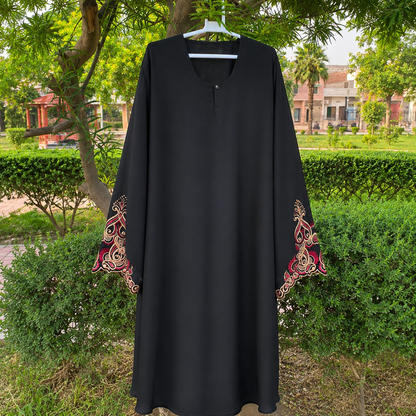 A black long-sleeve abaya dress with ornate red and gold embroidered patterns on the cuffs, hanging on a white hanger outdoors in front of green bushes and trees.