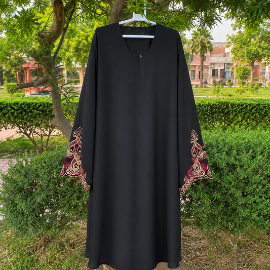 A black long-sleeve abaya dress with ornate red and gold embroidered patterns on the cuffs, hanging on a white hanger outdoors in front of green bushes and trees.