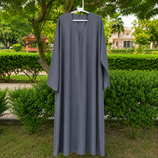 A long, loose-fitting gray abaya dress hanging on a white hanger outdoors in a garden setting with green hedges and trees.